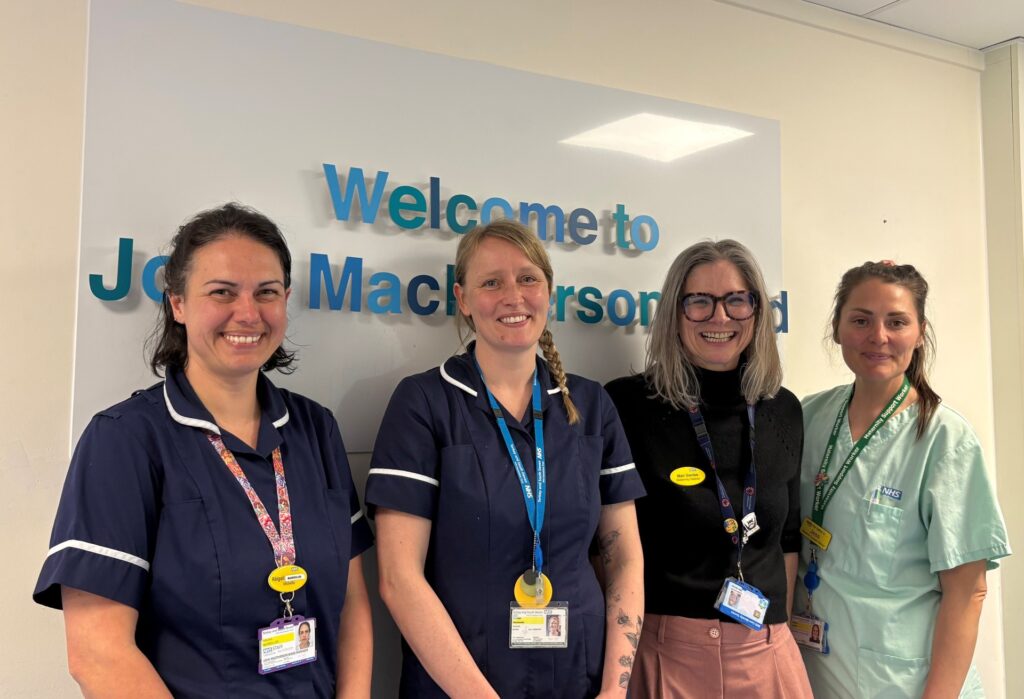 Four midwives standing in front of the Welcome to John MacPherson Ward sign