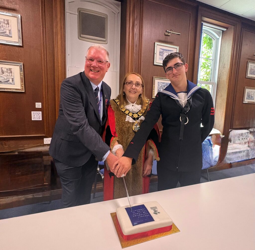 The Chair of the Torbay Royal Marines Association, , the Torbay Civic Mayor, and a Sea Cadet jointly cut a celebration cake at Torbay Hospital. The square white cake features a blue Armed Forces Covenant emblem and red ribbon bordering, celebrating the re-signing of the covenant.