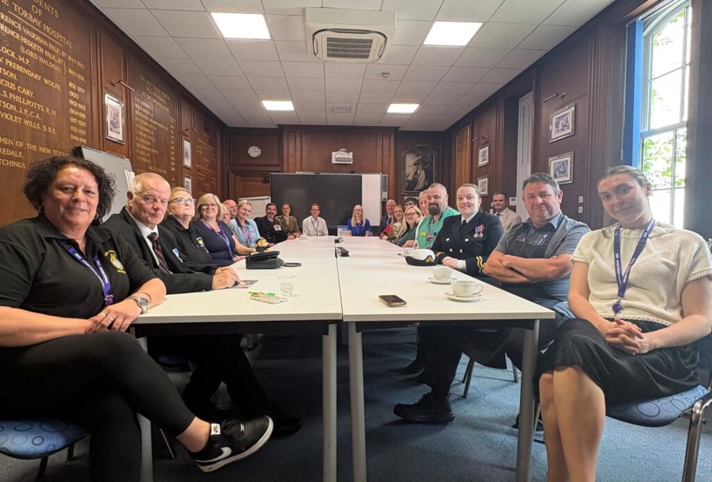 Representatives from across local communities—including Torbay and South Devon NHS Foundation Trust leadership, the Torbay Civic Mayor, members of the Torquay Armed Forces and Veterans Breakfast Club, Sea Cadets, and their Commanding Officer—gather around a long table in a wood-panelled room at Torbay Hospital to celebrate the re-signing of the Armed Forces Covenant.