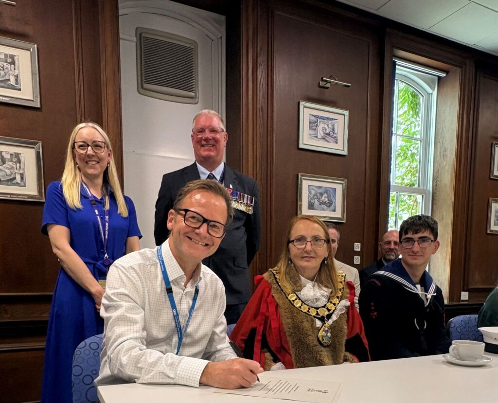 Joe Teape, Chief Executive of Torbay and South Devon NHS Foundation Trust, sits at a table writing on a document to re-sign the Armed Forces Covenant. Seated next to him is the Torbay Civic Mayor wearing official red and fur robes with a chain of office, and a Sea Cadet in uniform. Standing behind them are Jess Piper, Chief People Officer, and an Armed Forces representative wearing military medals.