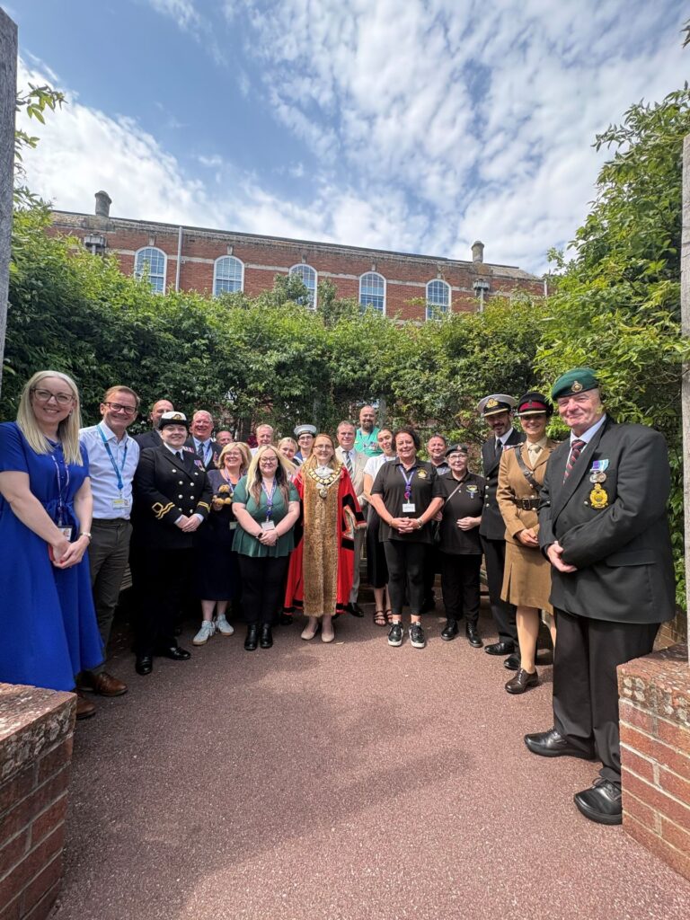 A group photo in a courtyard at Torbay Hospital featuring Jess Piper, Chief People Officer, Joe Teape, Chief Executive of Torbay and South Devon NHS Foundation Trust, the Torbay Civic Mayor, local veterans, Sea Cadets, and NHS staff posing together under a sunny sky to celebrate the re-signing of the Armed Forces Covenant.