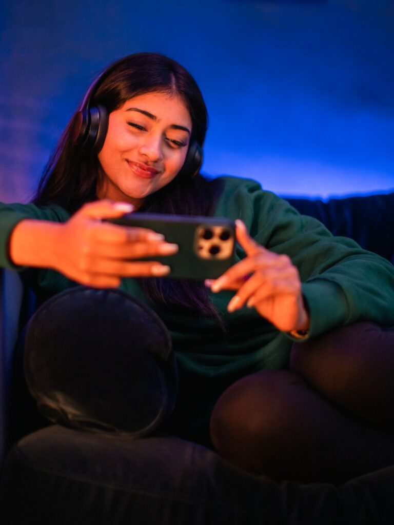 A young woman with long dark hair wearing black over-ear headphones sits comfortably on a couch while holding a dark-colored smartphone horizontally in both hands, smiling as she looks at the screen. The scene is illuminated with cozy, dual-tone ambient lighting—warm orange light on her right side and deep blue light on her left.