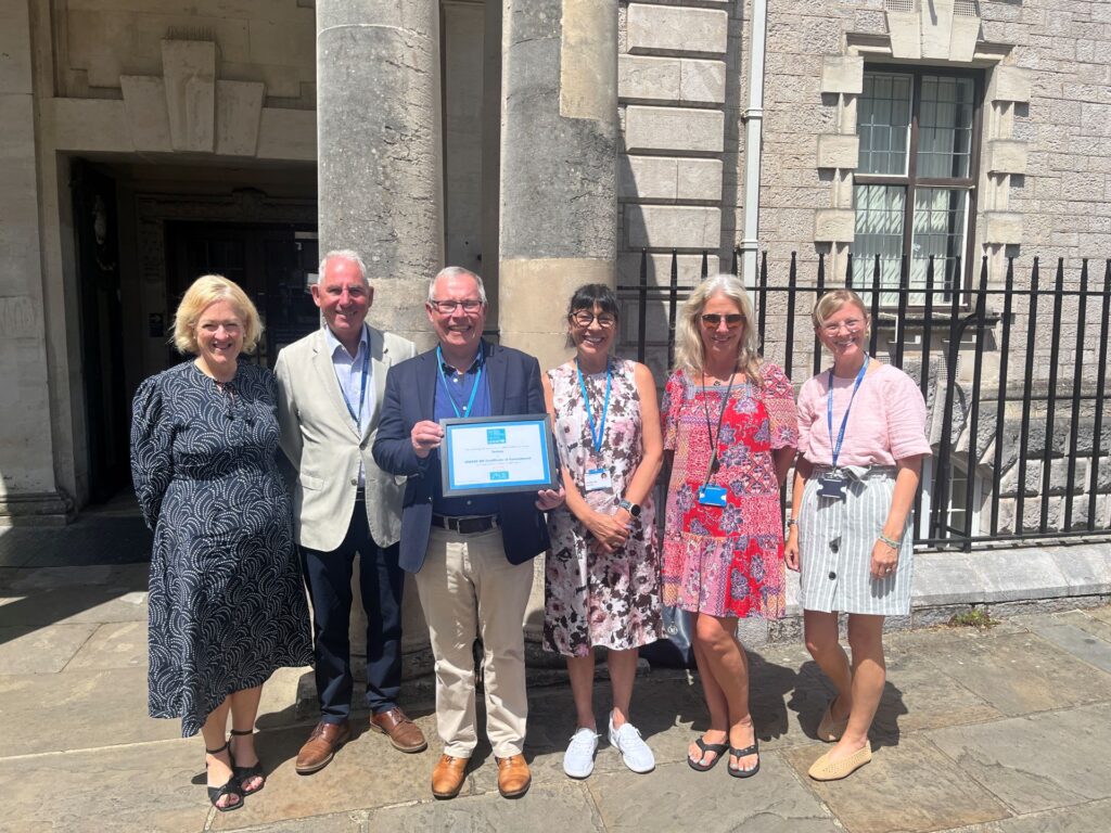 A group of six people standing outside a stone building with columns, holding a framed UNICEF UK Baby Friendly Initiative Certificate of Commitment.