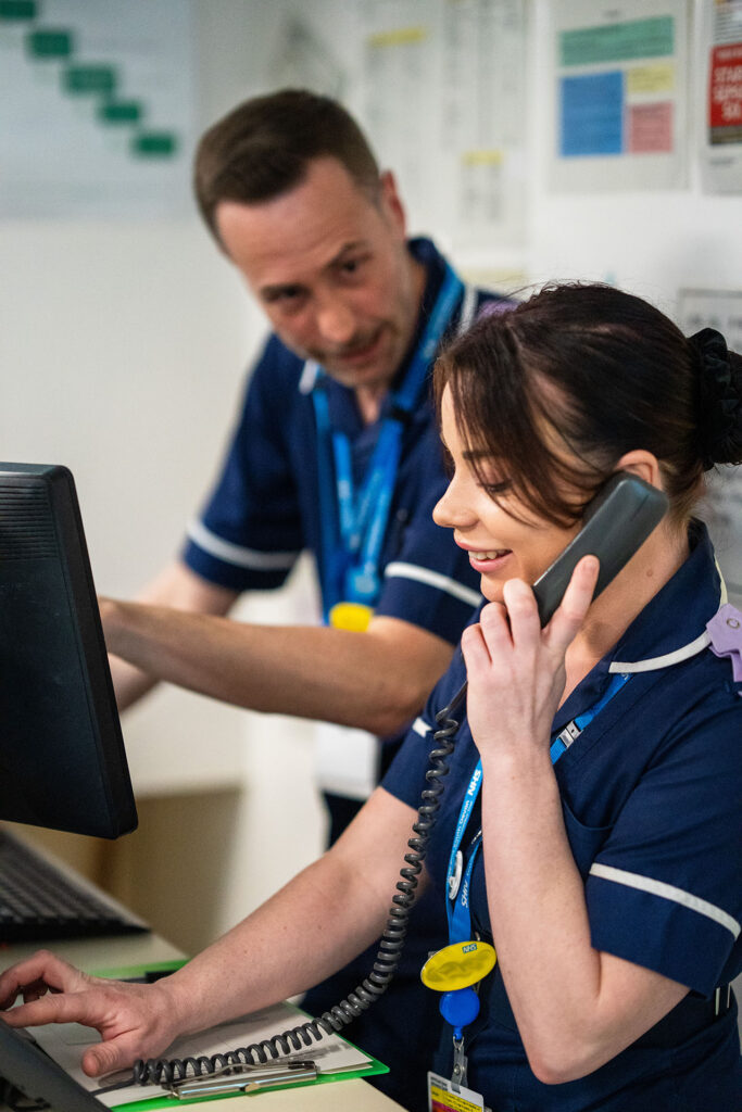 Two NHS colleagues at a reception desk with one on the phone