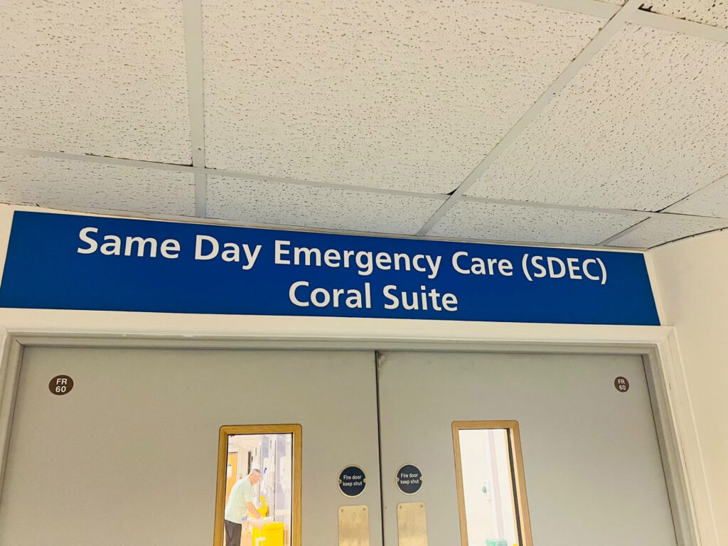 Double doors with vision panels beneath a blue sign that reads "Same Day Emergency Care (SDEC) Coral Suite" in white lettering.