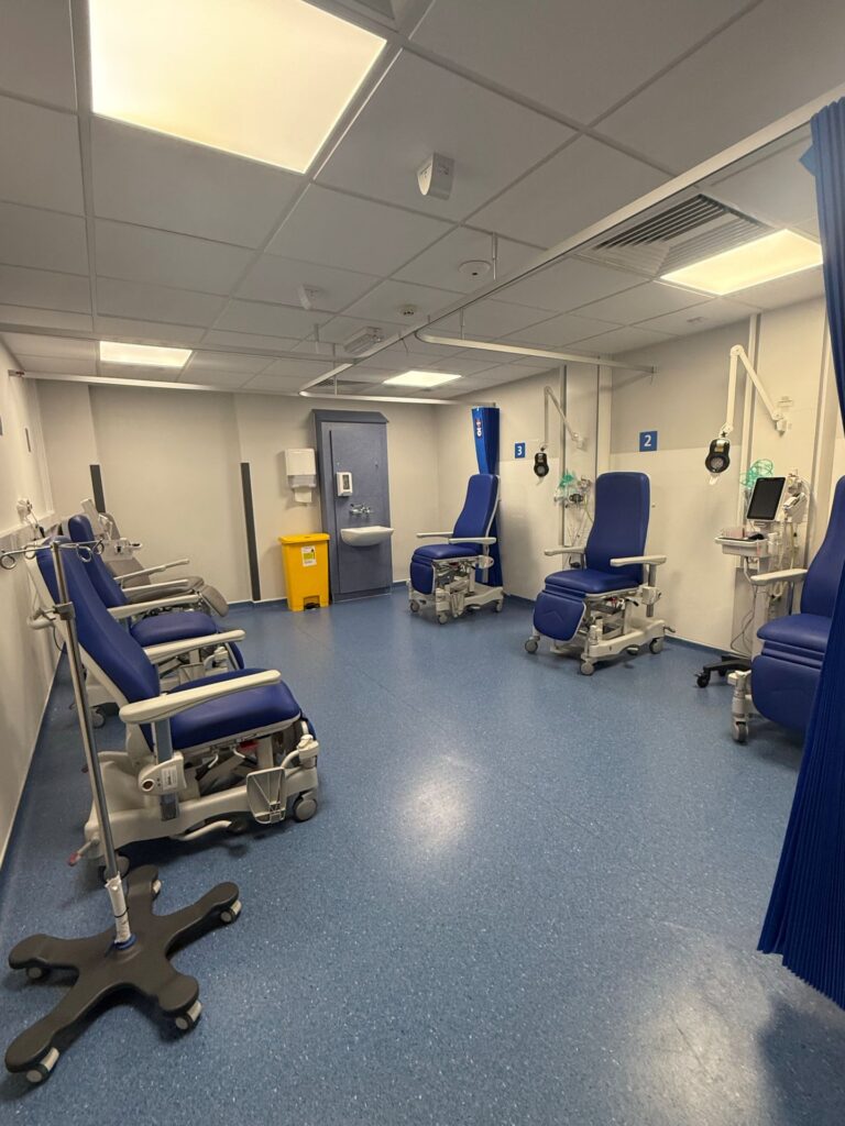 A clean hospital treatment bay featuring blue adjustable recliner chairs, IV poles, medical monitors, blue privacy curtains, and a handwashing sink unit.