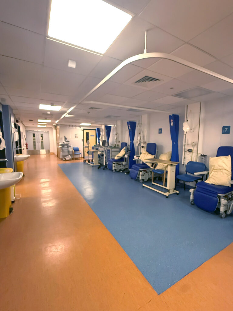 A wide hospital corridor with two-tone flooring featuring numbered patient bays with blue recliner chairs, yellow pillows, overbed tables, and blue privacy curtains.