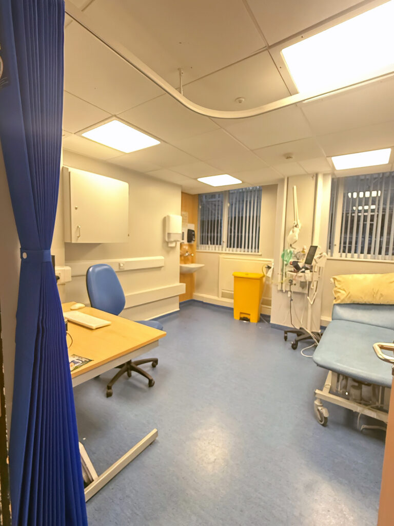 A clinical consultation room featuring a wooden desk with a computer and office chair, an examination couch with a pillow, medical equipment, and blue privacy curtains.